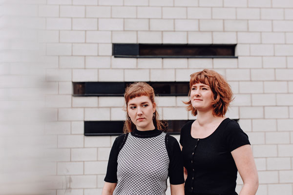 The Rheingans Sisters: Rowan and Anna against a white and black brick wall